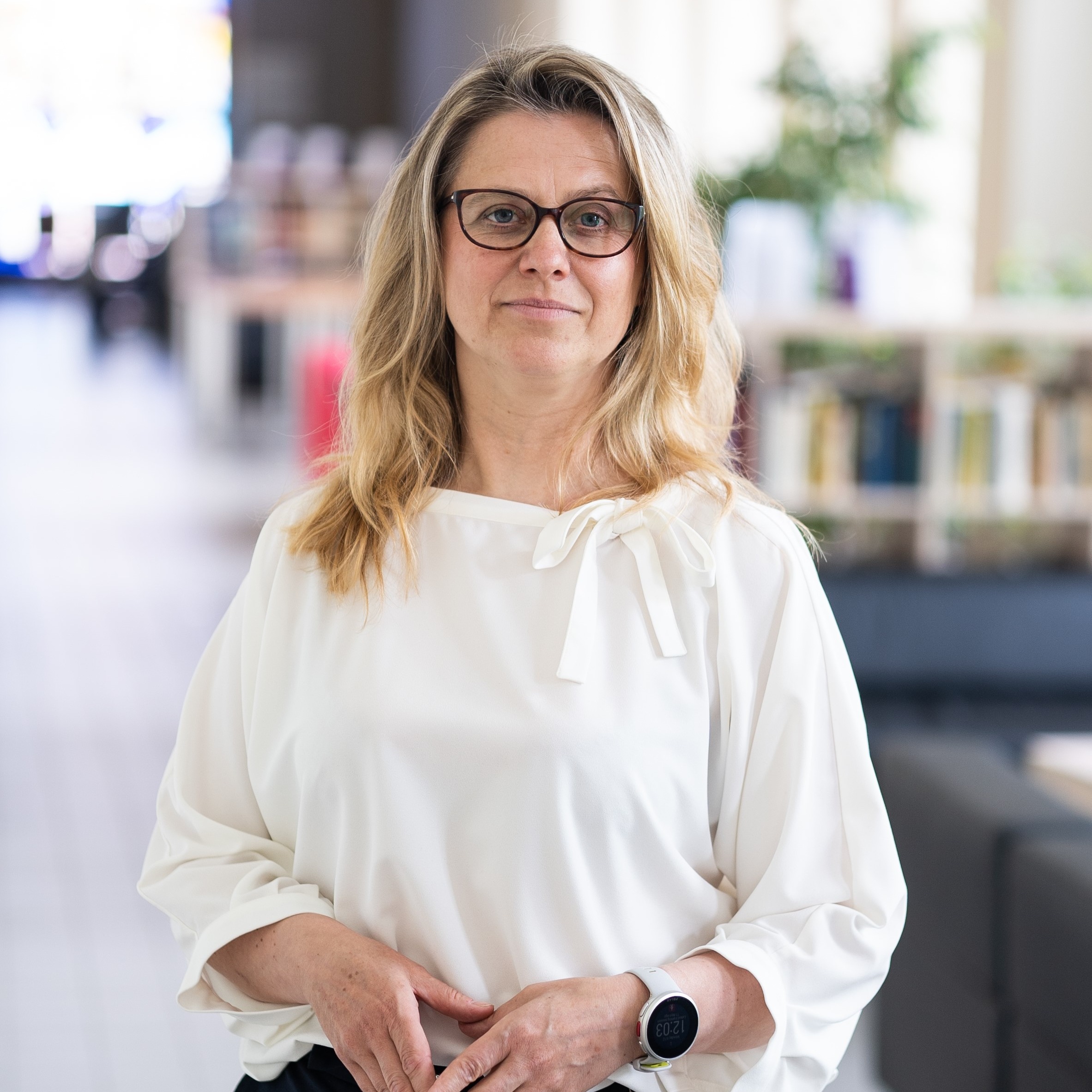 Dr. Laura Daniusevičiūtė-Brazaitė stovi moderniame interjere su knygomis fone, dėvinti baltą palaidinę ir laikanti rankas susikibus. Ji yra projekto vyresnioji mokslo darbuotoja, docentė.
