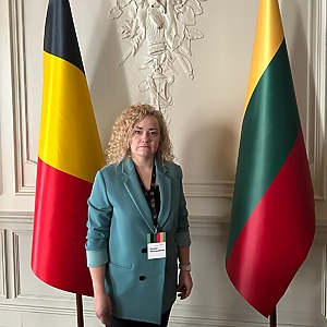 Dr. Donata Jovarauskienė stovi tarp Belgijos ir Lietuvos vėliavų, vilkinti melsvą &scaron;varką ir segėdama vardinę kortelę. Ji yra projekto vykdytoja, socialinių mokslų daktarė.