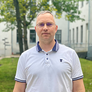 Dr. Rimantas Rauleckas stovi lauke prie universiteto pastato, dėvintis &scaron;viesiai pilkus mar&scaron;kinėlius. Jis yra projekto vyresnysis mokslo darbuotojas, docentas.